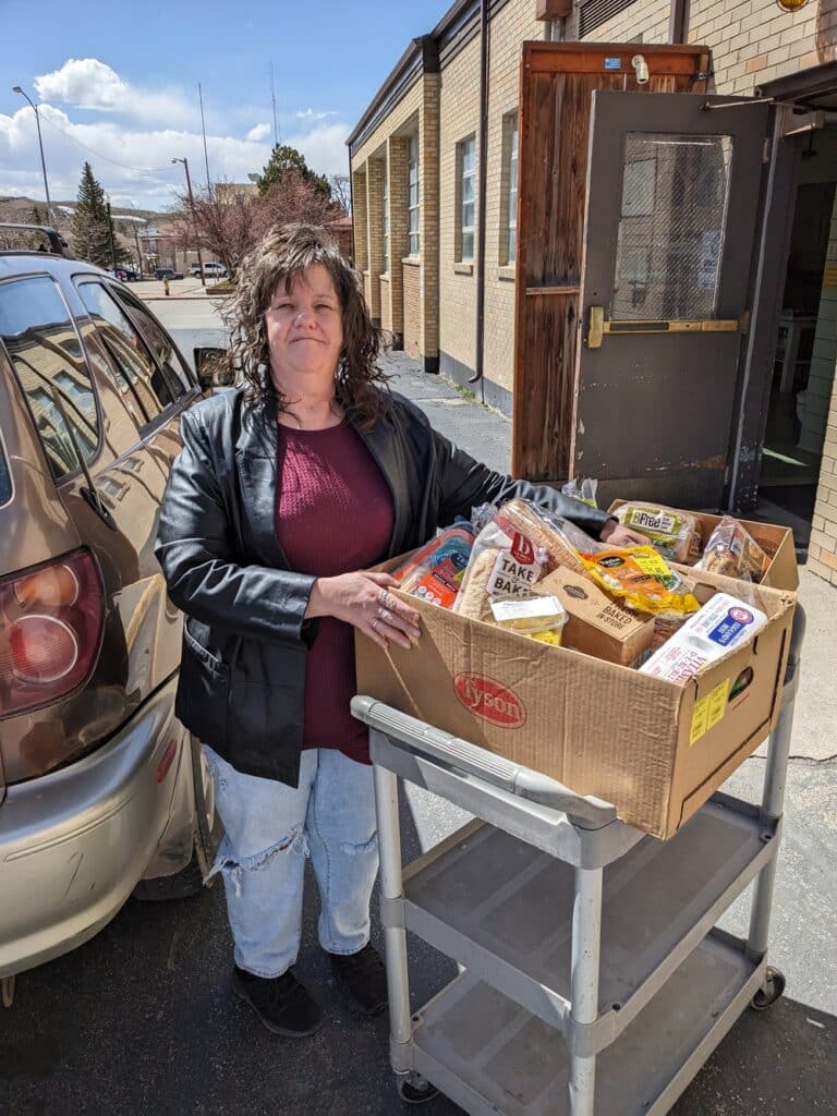 woman standing outside in between car and a cart with a box filled with food.