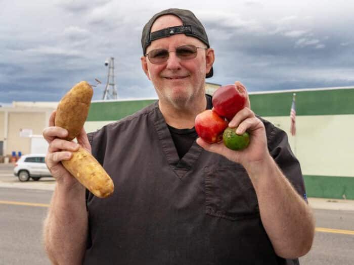 Man holding two potatoes in one hand and three peppers in his other hand, smiling.