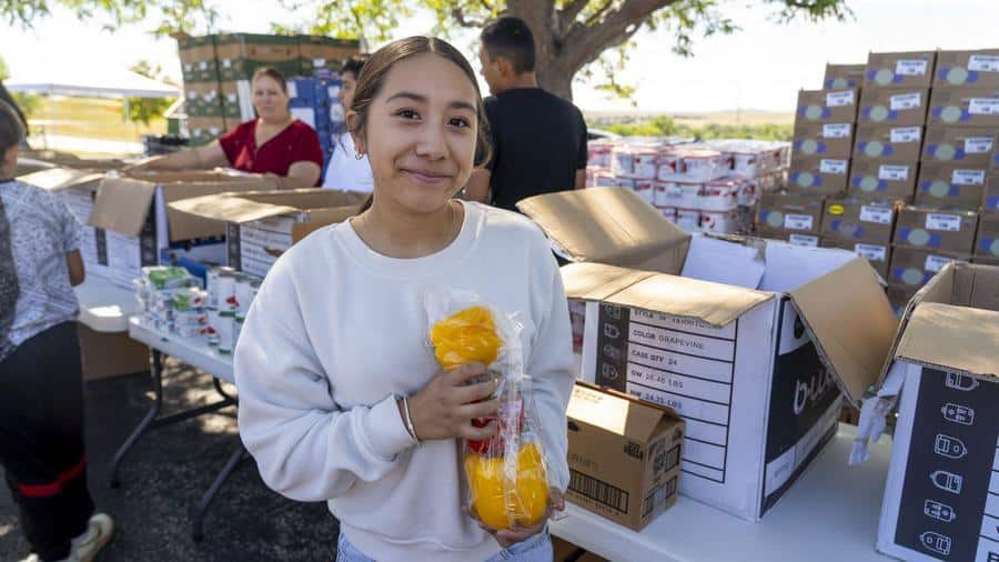 Woman at a food distribution holding a pack of yellow and red peppers, boxes and people in the background.