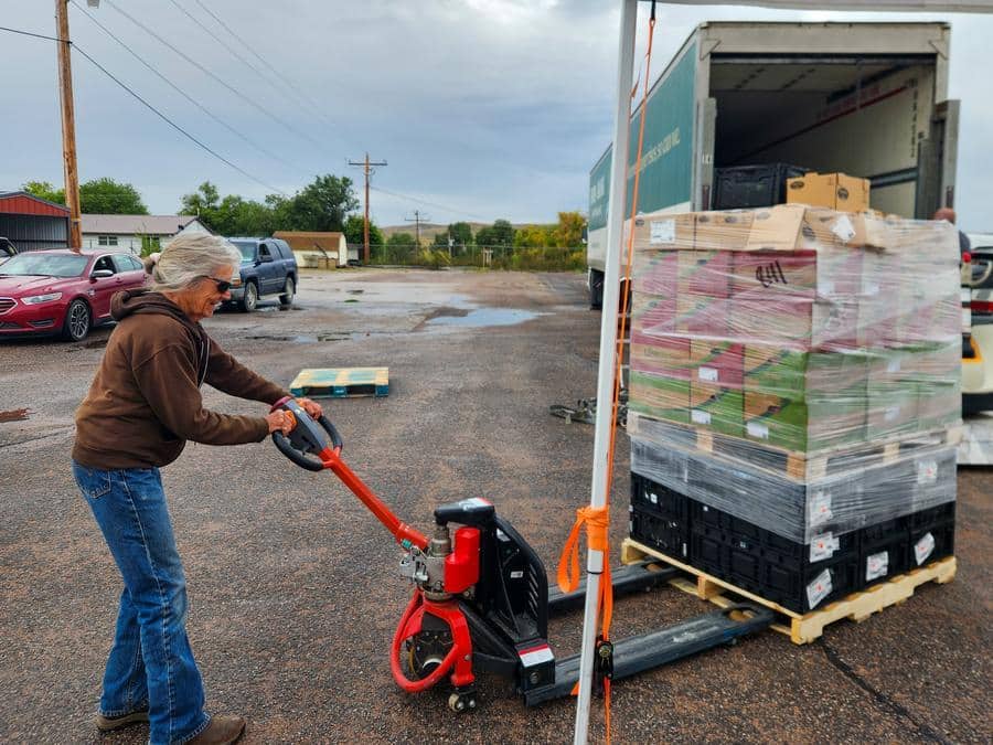 Woman using pallet jack to move a full pallet of fresh produce outside at a food distribution.