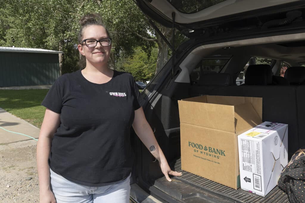 Woman with food box leaning against the trunk area of her car.