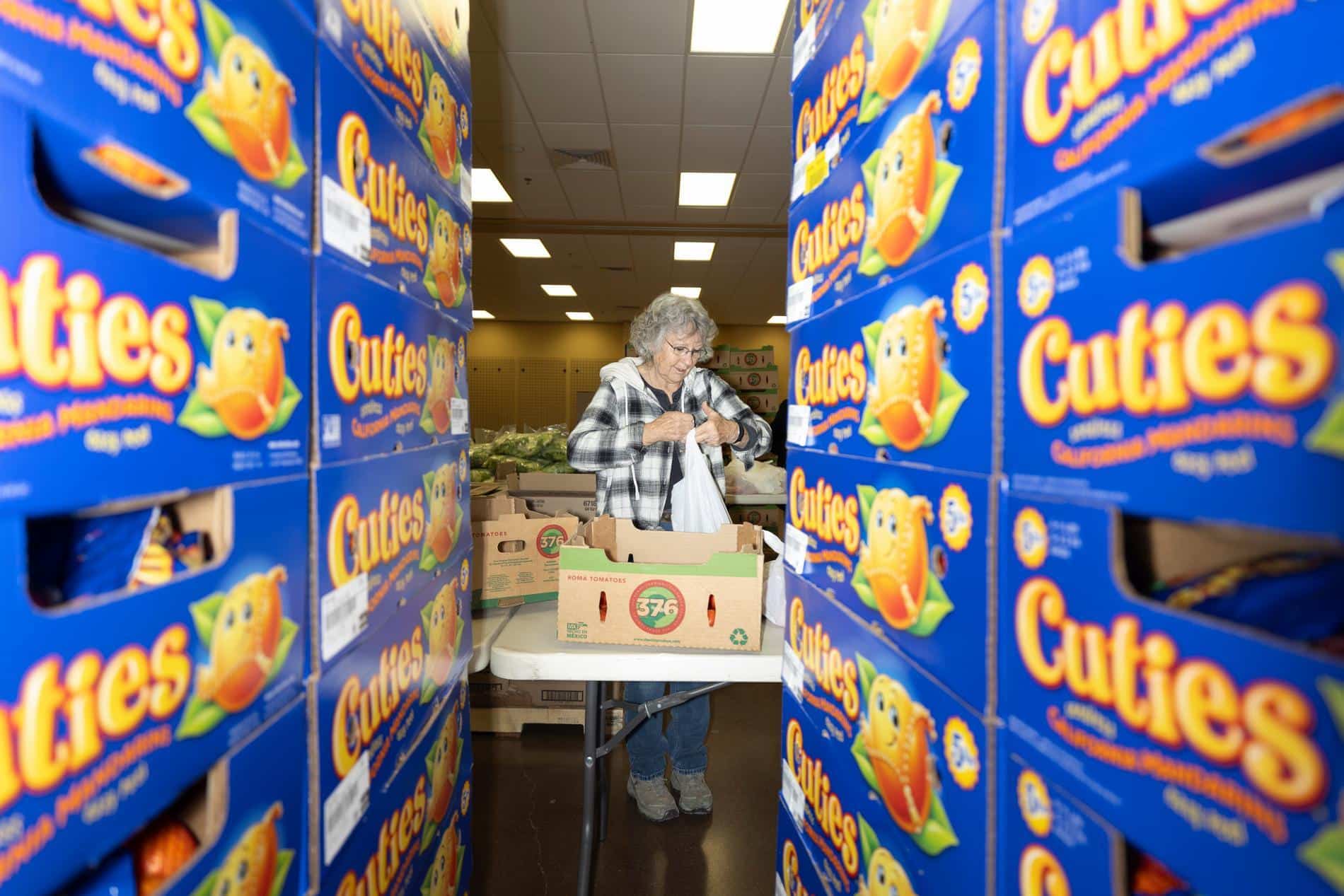 A volunteer in the background packing food into a bag. Foreground is boxes of Cuties stacked on left and right sides.