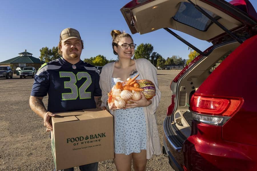 Couple standing behind car with trunk door open and holding a Food Bank of Wyoming box and a mesh bag of produce.